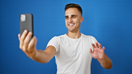 Smiling young hispanic man taking a selfie isolated against a vibrant blue background, radiating...