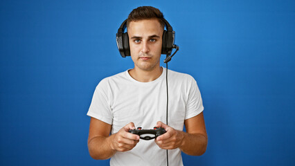 Handsome young man with headphones playing video games against a solid blue background.