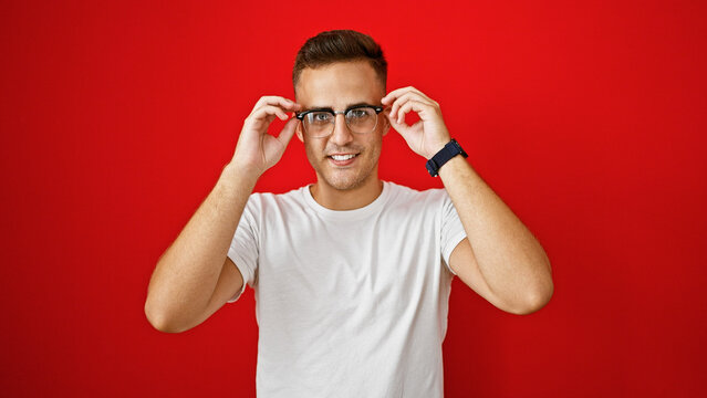 Handsome young hispanic man adjusting glasses against a vibrant red isolated background.