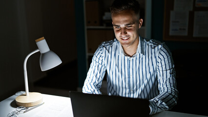 A focused young man works late on his laptop in a dimly lit office, emphasizing the modern work...