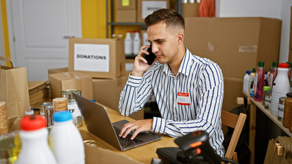 Handsome young man using laptop and phone at a donation center warehouse with food and supplies in background