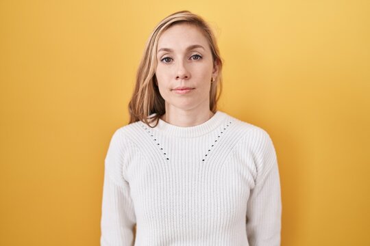 Young caucasian woman wearing white sweater over yellow background relaxed with serious expression on face. simple and natural looking at the camera.