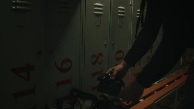 Medium tilting shot of young muscular professional male swimmer preparing for early morning practice in locker room in pool, getting towel, goggles and cap out of holdall bag, taking off hoodie