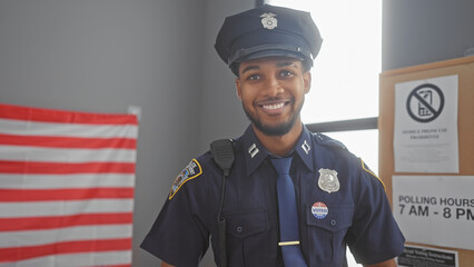 Smiling african american policeman with 'i voted' sticker in uniform posing in front of us flag indoors. - obrazy, fototapety, plakaty