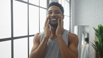 A smiling black man at home touches his beard, looking relaxed in a casual indoor setting.
