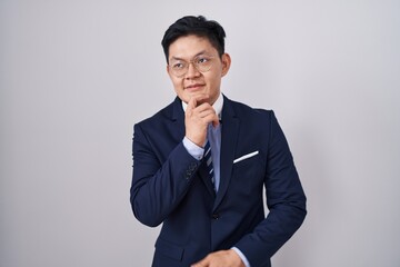 Young asian man wearing business suit and tie with hand on chin thinking about question, pensive expression. smiling and thoughtful face. doubt concept.