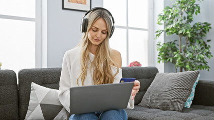 A blonde woman listens to headphones and holds a credit card while using a laptop at home,...
