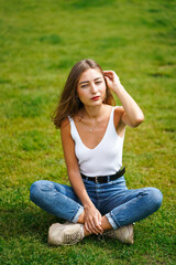 Fototapeta premium Beautiful young woman wearing tank top and jeans sitting on grass lawn