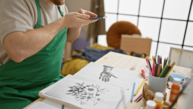 A young, bearded man in a studio captures his artwork with a smartphone, surrounded by creative tools and sketches.