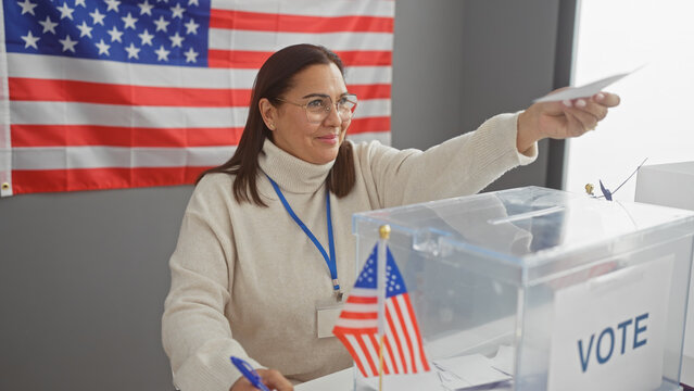 Mature hispanic woman election volunteer inserts ballot with us flag in background at voting center.