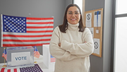 Confident hispanic woman smiles in a usa voting center with folded arms, american flag and ballot box