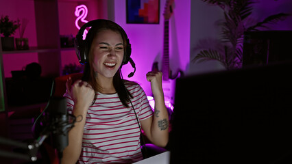 A joyful young brunette woman celebrates a victory in a neon-lit gaming room at night. © Krakenimages.com