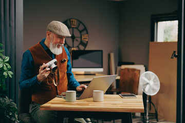 Portrait d'un homme photographe assis souriant quinquagénaire senior hipster élégant et stylé qui travaille sur un ordinateur et une tablette dans un atelier créatif vintage - 764952927