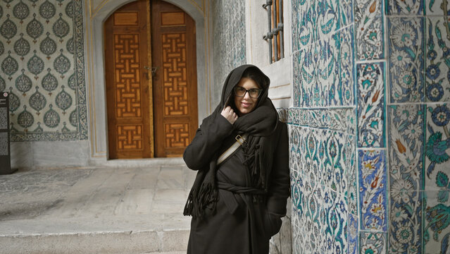 A young brunette woman wearing glasses and a shawl smiles by historic turkish tiles at topkapi palace, istanbul.