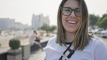 A smiling young adult woman wearing glasses, styled as a brunette, enjoys a sunny seaside cityscape...