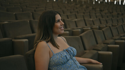 A young, smiling hispanic woman sits alone in a spacious theater, exuding a sense of leisure and...