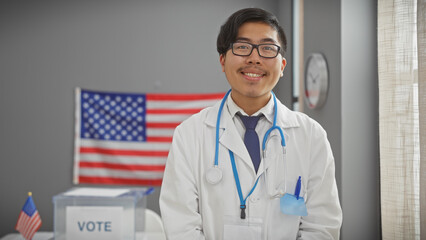 A young asian man with glasses, wearing a lab coat and stethoscope, smiles professionally in a us...