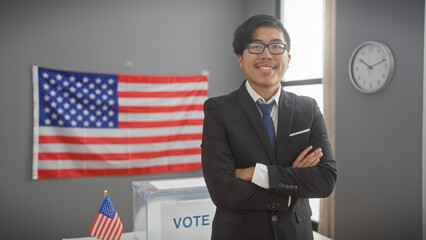 Smiling asian man with crossed arms standing in a modern interior with a us flag and a voting...