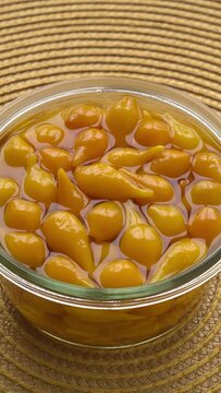 Pickled whole Biquinho Yellow chili in a glass jar on a yellow background. Sour and sweet Asian-style pickled homegrown chilies. Table spin, vertical video.