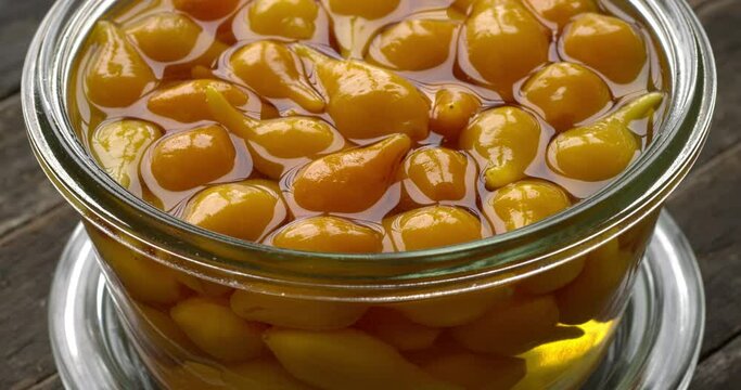 Close up view of pickled whole Biquinho Yellow chili in a glass jar on a rustic wooden table. Sour and sweet Asian-style pickled homegrown chilies. Table spin.