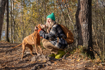 Middle aged happy woman petting vizsla dog walking in nature autumn forest. Female rejoicing spending time with pet friend on weekend sitting on haunches.