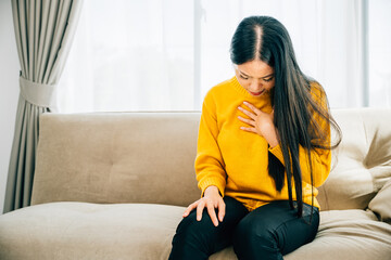 Portraying a heart attack, Asian woman on sofa holds her left chest in pain requiring immediate medical assistance. Illustrating heartache discomfort and the need for emergency aid. - obrazy, fototapety, plakaty