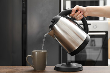 Woman pouring hot water from electric kettle into cup in kitchen, closeup