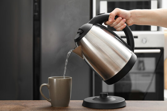 Woman pouring hot water from electric kettle into cup in kitchen, closeup
