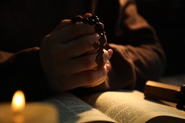 Woman praying at table with burning candle and Bible, closeup - obrazy, fototapety, plakaty