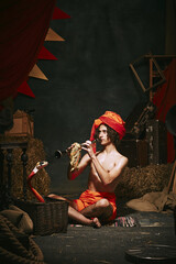 Young man, charmer in turban playing flute with cobra emerging from basket over dark retro circus...