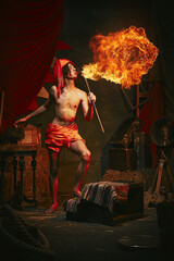 Fire show. Young man in turban breathes fire, displaying dramatic flame burst over dark retro circus backstage background. Concept of circus, theater, performance, show, retro and vintage © master1305
