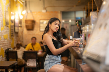 A young woman is sipping a cold alcoholic drink. While sitting at the bar in a rustic cafe and looking at camera.