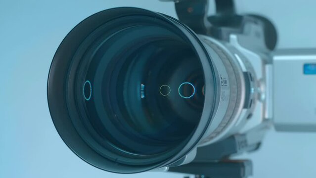 In the studio, ring LED light is reflected in a powerful zoom lens of a professional video camera standing on a tripod against a light background. Closeup. Macro. Shot in motion