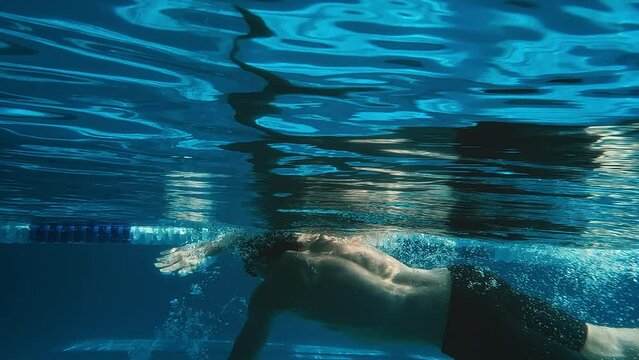 Full slowmo underwater footage of unrecognizable male athlete or customer in black trunks, cap and goggles swimming freestyle in lane at pool