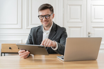 A cheerful businessman in a suit works on a tablet with a laptop open on the desk in a classic...