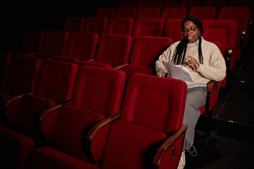 Minimal portrait of Black adult woman reading script in audience at theater in low light copy space