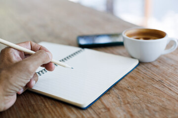 Select focus on hand using pencil taking note in book with coffee cup and mobile on old wooden table