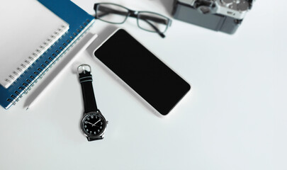 Top view, wrist watch with mobile phone, notebook, eyeglasses and digital camera on white table