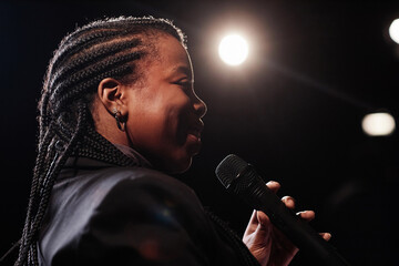 Side view portrait of smiling Black woman holding microphone and performing on stage with spotlight...