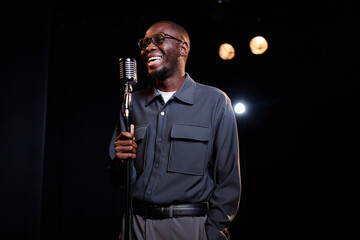 Waist up portrait of smiling young Black man speaking to microphone on stage while performing in...