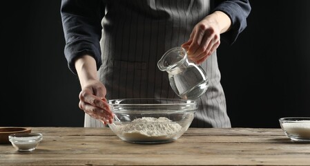 Making bread. Woman pouring water into bowl with flour at wooden table, closeup