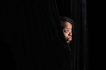 Young African American woman peeking from curtains looking at theater stage, copy space