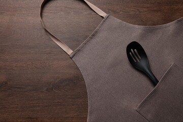 Stylish brown apron and slotted spoon on wooden table, top view. Space for text