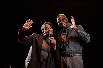 Portrait of two Black performers on stage speaking to microphone and waving hello greeting audience