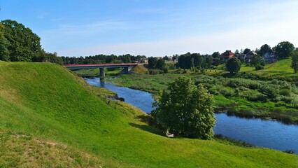 A small town with houses and gardens stands on the riverbank slope. A bridge on concrete piers with metal fencing and light poles connects the banks of the river. Sunny summer weather and blue sky