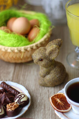 Easter eggs in the basket, bowl of cookies, chocolate pralines, Easter bunny figurine, cups of tea, glasses of juice, flowers and lit candles on the table. Selective focus.