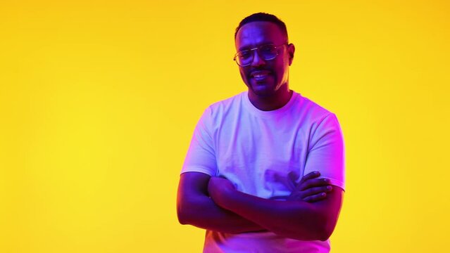 Confident young African-American man in glasses, white t-shirt posing, standing against yellow studio background in neon light. Concept of human emotions, casual fashion, lifestyle