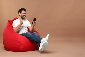 Happy man with smartphone sitting on bean bag against light brown background. Space for text