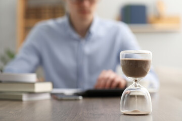 Hourglass with flowing sand on desk. Man using calculator indoors, selective focus