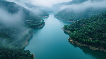Fog envelops the hills surrounding a tranquil river in a gorge, with verdant green foliage accentuating the peaceful scene.